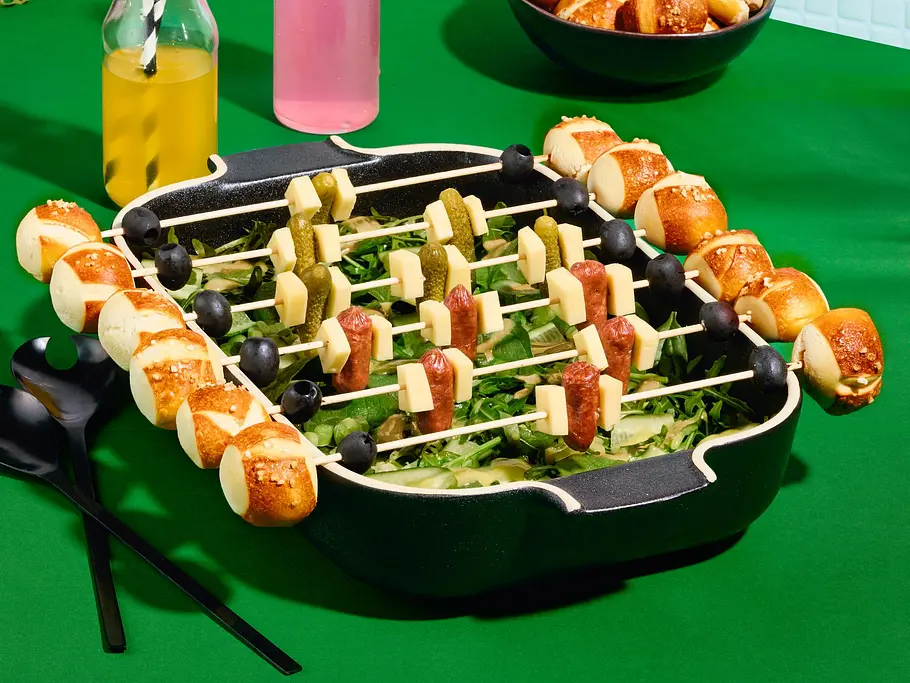 Salat in einer Auflaufform, darauf Laugenspieße mit Mini-Laugenbrötchen, Käsewürfeln, Oliven, Würstchen und Cornichons.