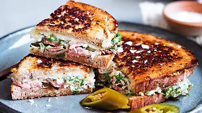 Geröstete Sandwichhälften mit Käse, Fleisch und Kräutern liegen neben grünen Jalapenostücken auf einem grauen Teller. - Foto: LECKER @ Bauer Media Group Geröstete Sandwichhälften mit Käse, Fleisch und Kräutern liegen neben grünen Jalapenostücken auf einem grauen Teller. - Foto: LECKER @ Bauer Media Group