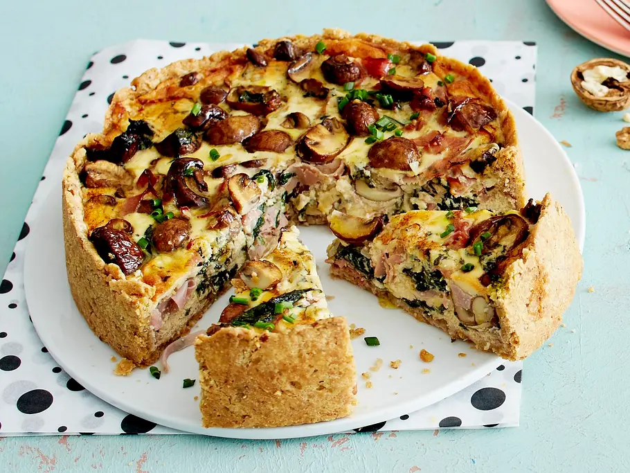 Schinken-Käse-Quiche mit Champignons Rezept Auf einem hellblauen Tisch mit gepunkteter Serviette steht eine angeschnittene überbackene Pilz-Quiche mit sichtbaren Schinkenstreifen und Spinatgrün im Anschnitt.