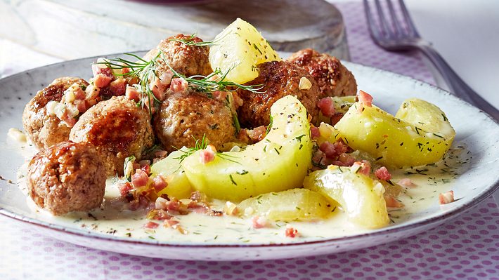 Die Schmorgurken liegen in cremiger Soße auf einem hellen Teller im Vordergrund. Dahinter sind Hackbällchen zu sehen. - Foto: House of Food / Bauer Food Experts KG