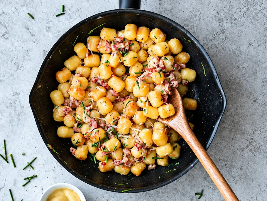 Schnelle Flammkuchen-Gnocchipfanne Gnocchi mit Speck, Käse und Schnittlauch liegen in einer schwarzen Pfanne mit Holzlöffel. Daneben steht eine Schale Senf.