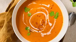 Cremige Tomatensuppe mit Kokosmilch, garniert mit frischem Koriander und einem Swirl aus Kokosmilch, serviert in einer weißen Schale auf einem hellen Holztisch mit gelbem Stoffserviette. - Foto: LECKER @ Bauer Media Group