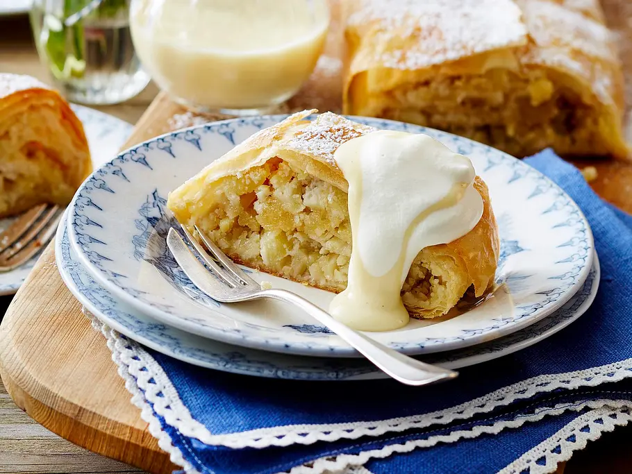 Ein Stück Apfelstrudel mit Vanillesoße auf einem Teller, daneben eine Gabel, im Hintergrund weiterer Strudel.