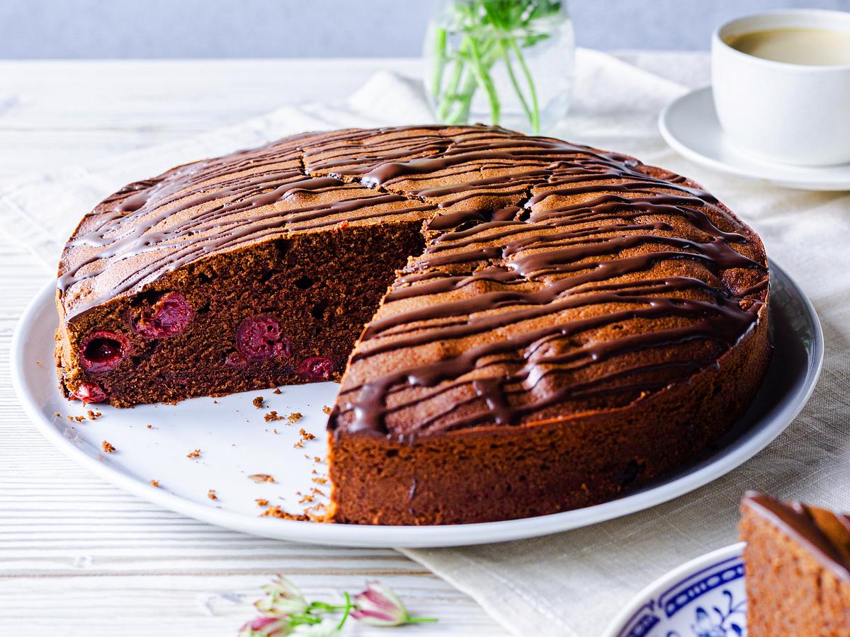 Der in einer Springform gebackene Schoko-Kirsch-Kuchen liegt auf einem Kuchenteller. Ein großes Stück ist herausgeschnitten, sodass man die Kirschen im dunklen Teig erkennt.