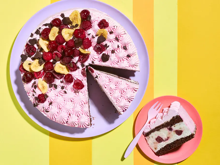 Schokoladige Kiba-Torte Eine rosa verzierte Torte mit Früchten und Schokolade steht neben einem Stück auf einem Teller auf gelb gestreiftem Grund.