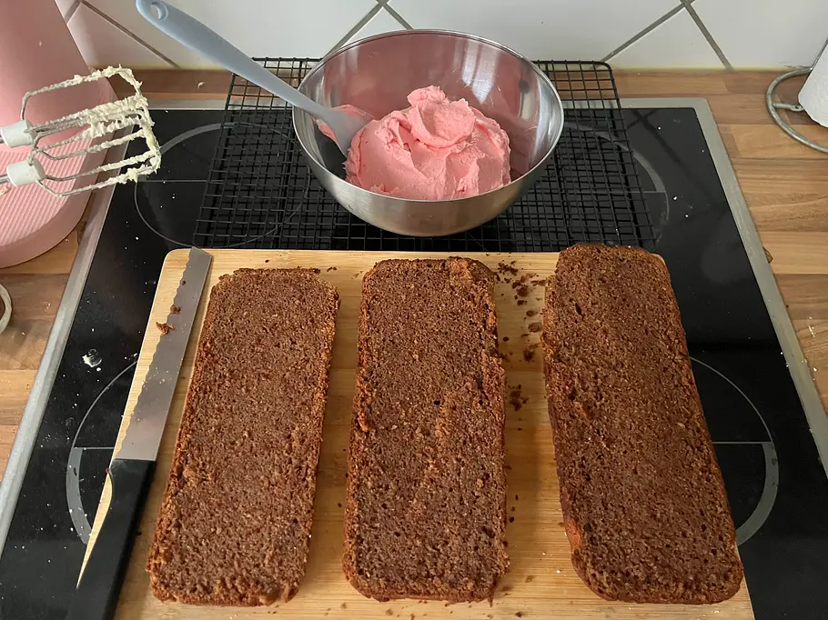 3 rechteckige Kuchenplatten in Form einer Kastenform auf einem Holzbrett, im HIntergrund rosa Buttercreme.