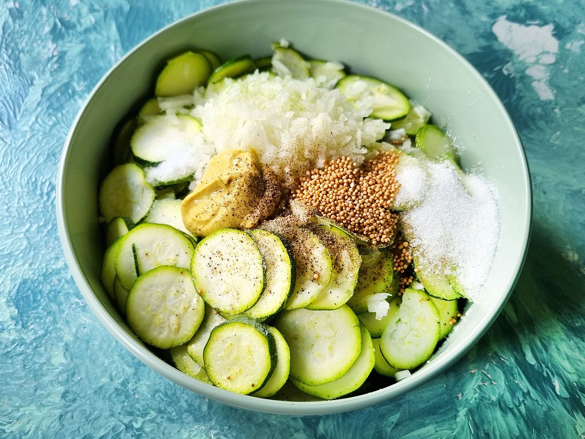 Zucchinischeiben, Senfkörner, Zwiebelwürfel, Zucker und Salz in einer Schale