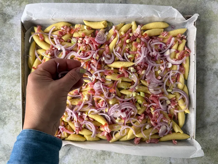 Eine Hand streut Frühlingszwiebeln über ein Blech mit Schupfnudeln, roten Zwiebelringen und Speckstückchen.
