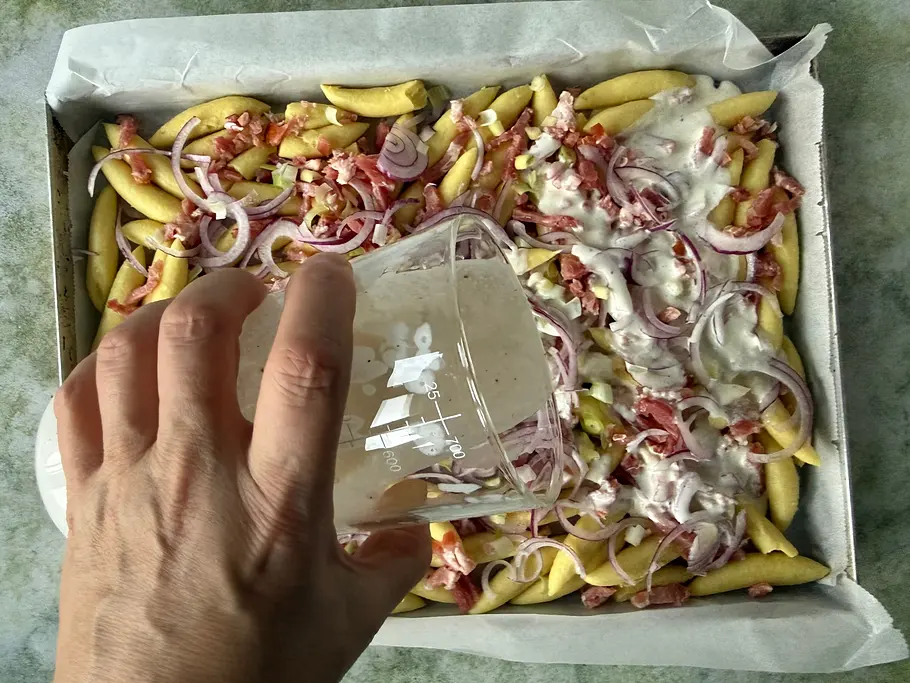 Eine Hand gießt helle Soße über Schupfnudeln mit roten Zwiebelringen und Speckwürfeln auf einem mit Backpapier belegten Blech.