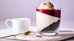Schwarzwälder Kirsch-Pannacotta im Glas mit weißer Creme, dunklem Schokoboden, Kirschspiegel und Sahnetupf, serviert auf heller Platte mit einer weißen Tasse und goldenem Löffel. - Foto: LECKER @ Bauer Media Group