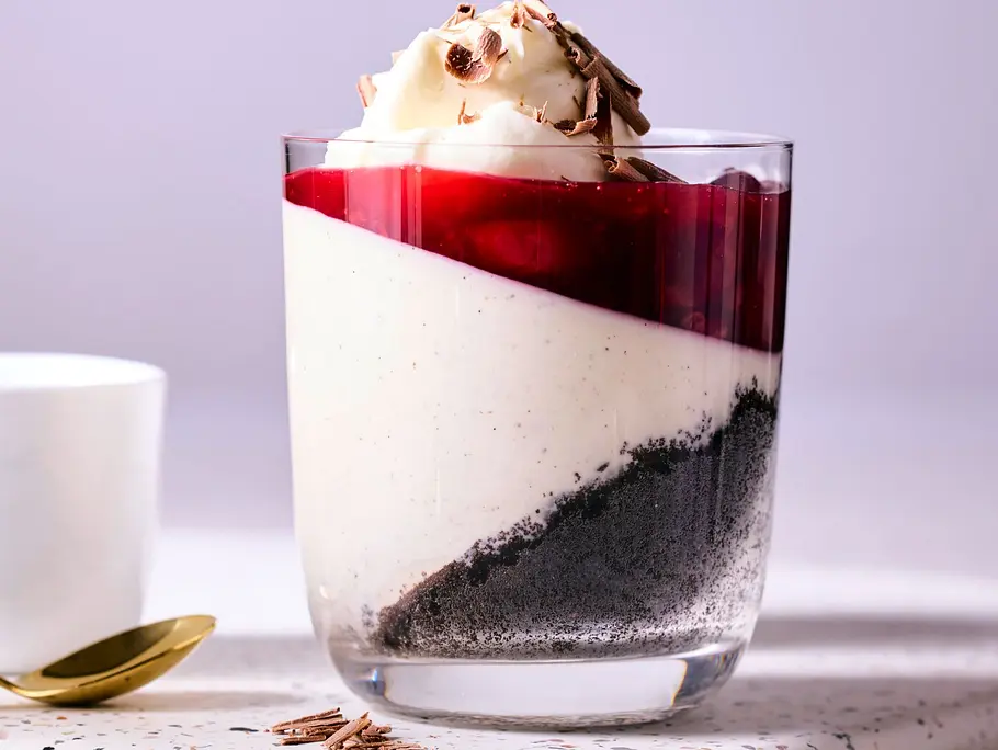 Schwarzwälder Kirsch-Pannacotta im Glas mit weißer Creme, dunklem Schokoboden, Kirschspiegel und Sahnetupf, serviert auf heller Platte mit einer weißen Tasse und goldenem Löffel.