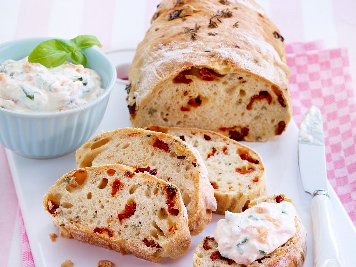 Selbst gebackenes Brot mit getrockneten Tomaten, in Scheiben geschnitten und serviert mit einem cremigen Dip in einer Schale - angerichtet  mit einem  rosa Geschirrtuch.
