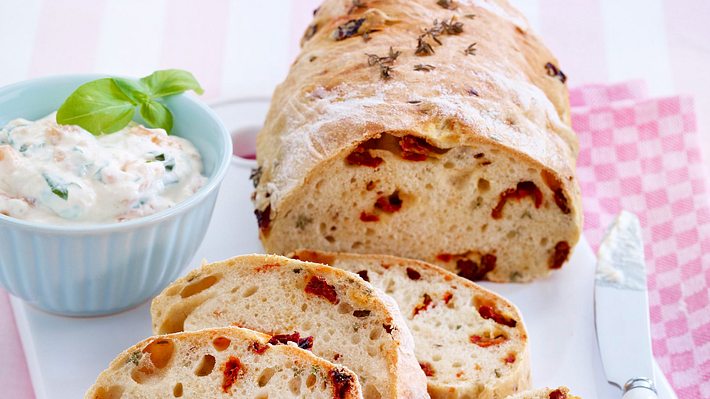 Selbst gebackenes Brot mit getrockneten Tomaten, in Scheiben geschnitten und serviert mit einem cremigen Dip in einer Schale - angerichtet  mit einem  rosa Geschirrtuch. - Foto: House of Food / Bauer Food Experts KG