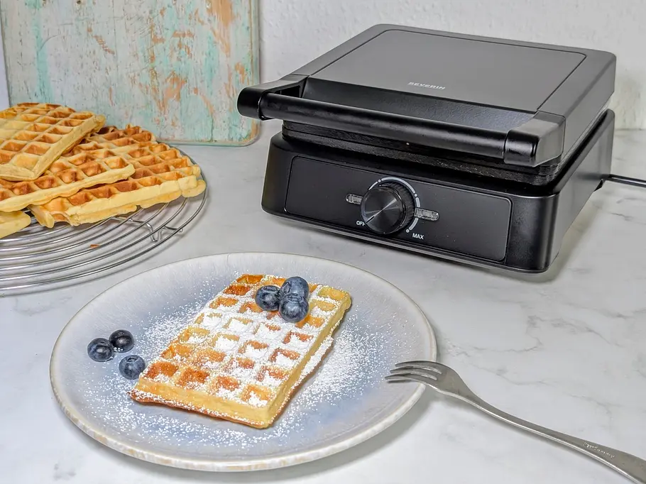 Das Severin WA 2130 Waffeleisen im Test Das Severin WA 2130 Waffeleisen steht geschlossen auf einer Arbeitsfläche, schräg von vorne zu sehen. Davor ein Teller mit einer darin gebackenen Belgischen Waffel, samt Puderzucker und Blaubeeren.