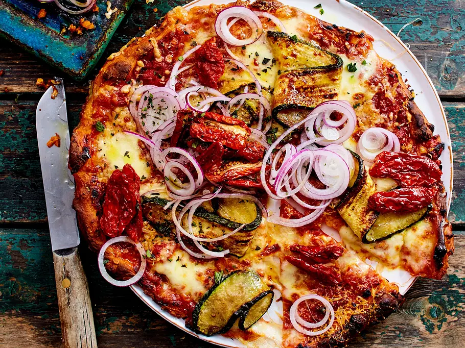 Süßkartoffel-Pizza mit Zucchini und Zwiebeln Eine Pizza mit Zucchini, Tomaten, Käse und roten Zwiebelringen liegt neben einem Messer und zwei Flaschen auf einem Holztisch.