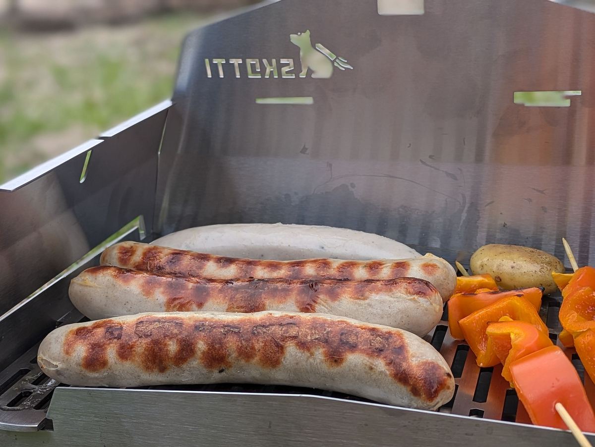 Skotti Gasgrill Brandings Auf dem Skotti Grill brutzeln drei Würstchen, eine Bratwurst, Paprikaspieße und eine kleine Kartoffel.