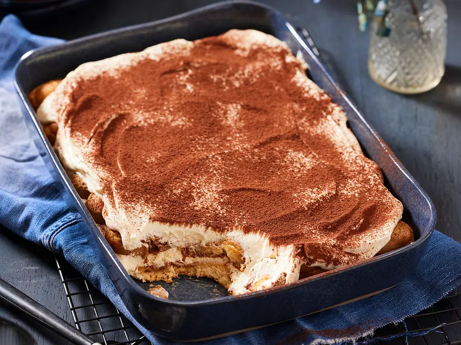 Skyr-Tiramisu in einer rechteckigen Auflaufform, mit Kakaopulver bestäubt, eine Portion ist bereits herausgenommen; serviert auf einem Tisch.