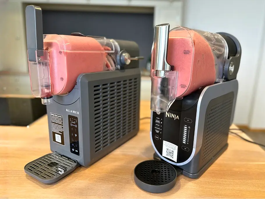 Die besten Slush-Maschinen im Test Die Slush-Maschinen von Klamer und Ninja stehen im Vergleich auf einer hölzernen Kücheninsel und haben beide rotes Slush-Eis im transparenten Behälter.