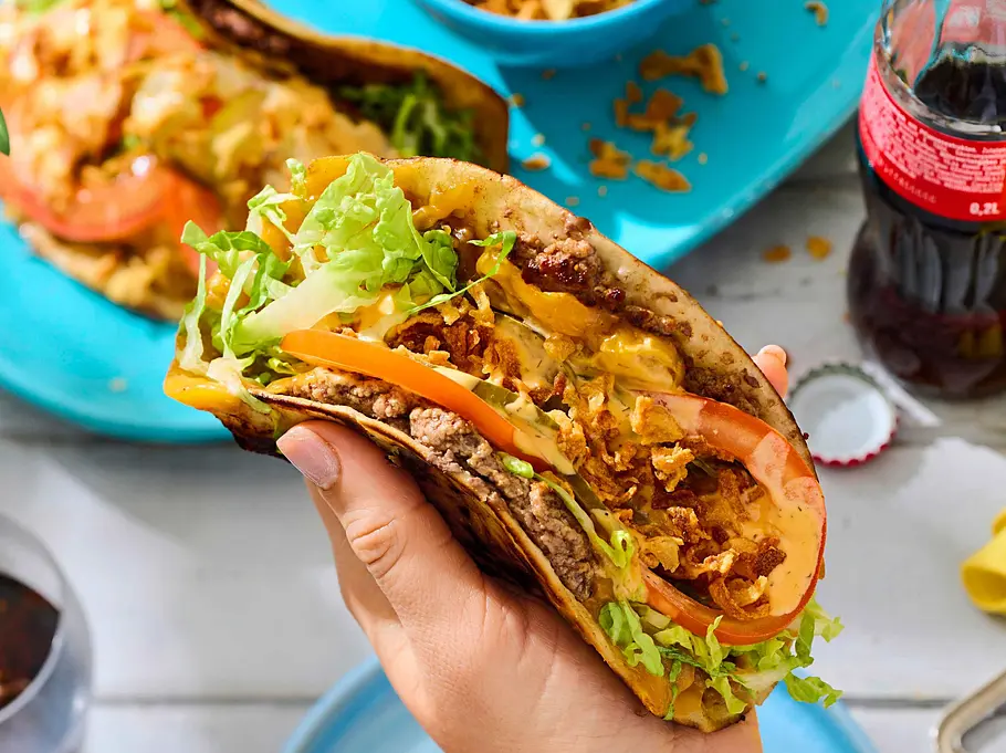 Smashed Cheeseburger-Tacos Rezept Smashed Cheeseburger-Tacos: Ein Taco geformt in einer Hand, gefüllt mit Hackfleisch, Salat, Tomate, geschmolzenem Cheddar, sauren Gurken und einer Burgersoße.