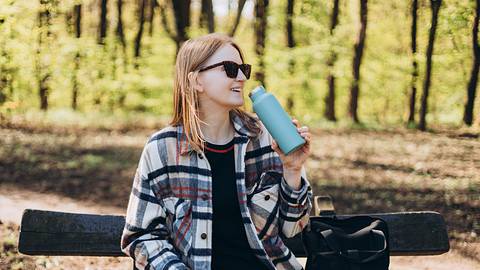 Frau mit Sonnenbrille sitzt auf einer Bank im Park und trinkt aus einem Thermobecher - Foto: iStock / Mariia Skovpen