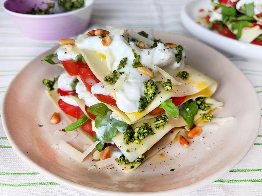 Sommer-Lasagne ohne Ofen Rezept Geschichtete Lasagneplatten auf einem Teller: gefüllt mit Tomaten, grünem Pesto, Rucola, Burrata; mit Pinienkernen, Parmesan und Pfeffer verziert.