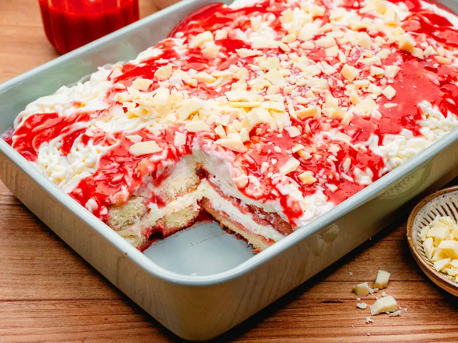 Spaghettieis-Tiramisu Rezept Eine blaue Auflaufform, darin ein geschichtetes Tiramisu abwechselnd weiße und rote Creme dekoriert mit Raspeln aus weißer Schokolade. Ein Stück vorne fehlt.