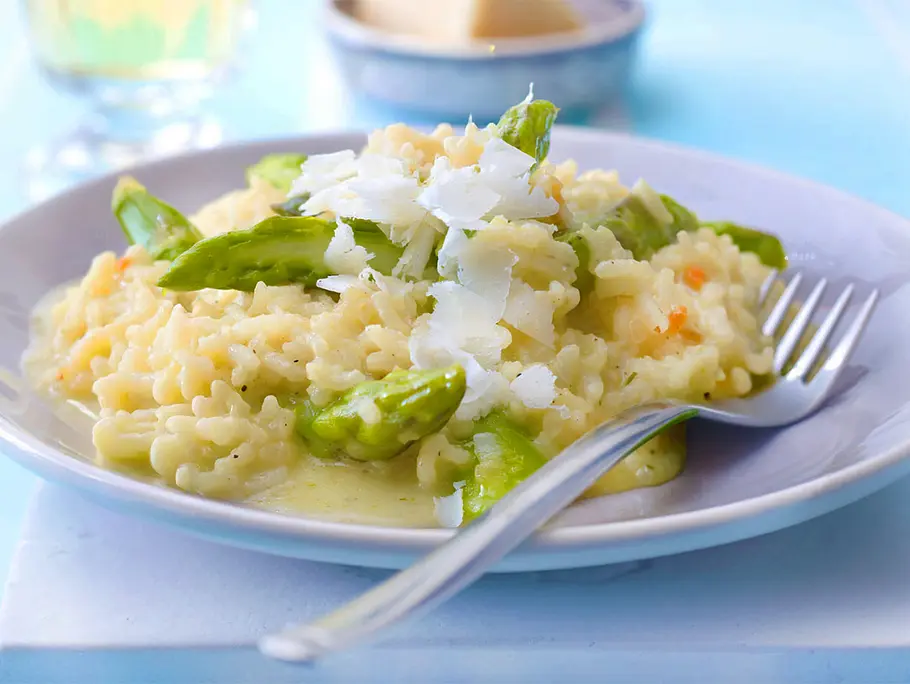 Spargelrisotto mit grünem Spargel Eine Portion Spargelrisotto mit grünem Spargel und Parmesan liegt auf einem Teller, im Hintergrund ein Schälchen mit Parmesan und ein Glas Weißwein