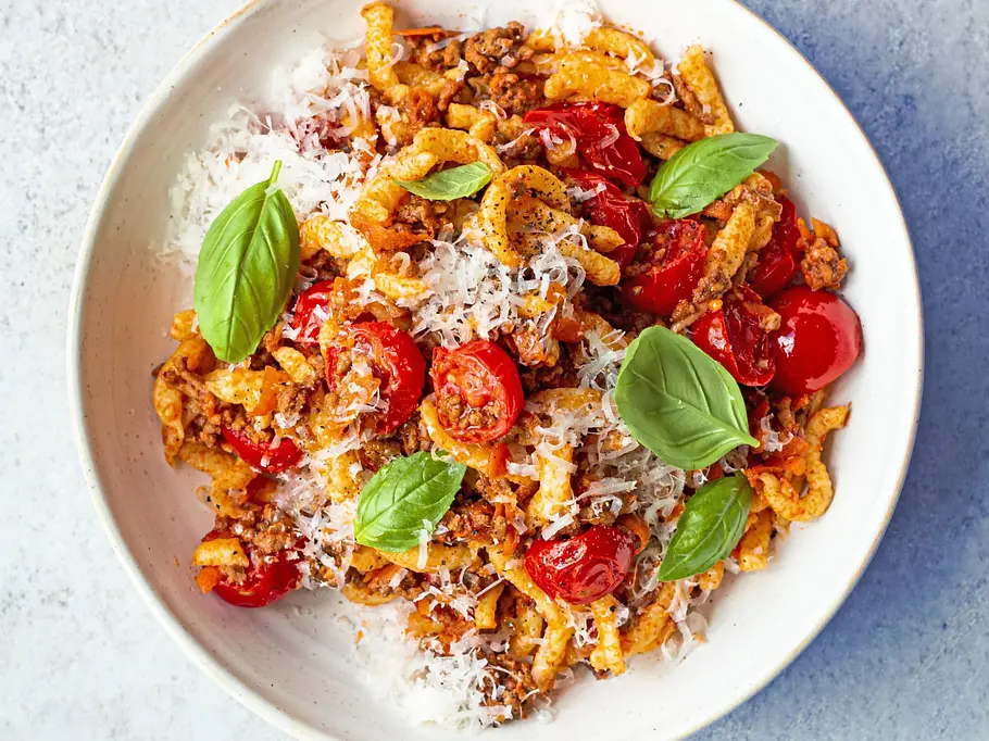 Spätzle mit Blitz-Bolognese Spätzle mit Hackfleisch-Bolognese, Kirschtomaten und frisch geriebenem Parmesan, garniert mit Basilikum, angerichtet auf einem weißen Teller.