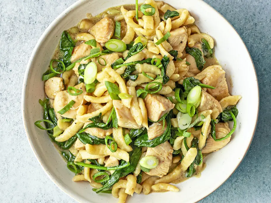 Spätzle mit zarten Hähnchenstücken, grünem Curry und frischem Blattspinat, garniert mit Frühlingszwiebeln, serviert auf einem weißen Teller.