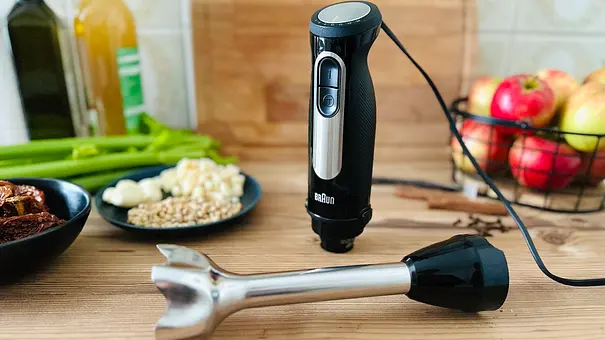 Ein abgeschraubter Stabmixer mit Pürierstab steht auf einer hölzernen Arbeitsfläche neben einer Schale mit Äpfeln, Stangensellerie und Zutaten für Pesto - Foto: LECKER @ Bauer Media Group / Nicole Molitor
