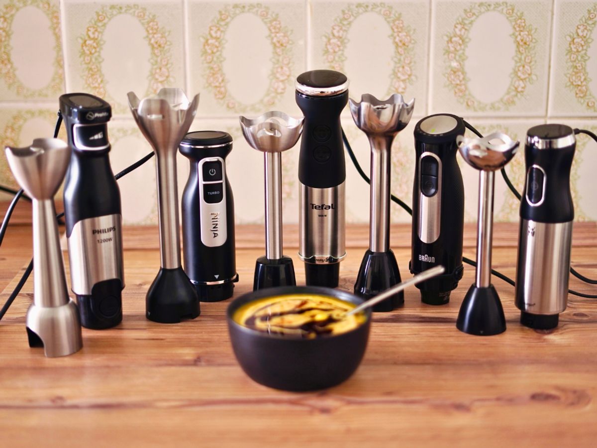 Stabmixer im Vergleich getestet Fünf verschiedene Stabmixer mit abgenommenem Mixtstab stehen nebeneinander vor einer Schale mit gelbe Kürbissuppe auf einer hölzernen Arbeitsplatte.
