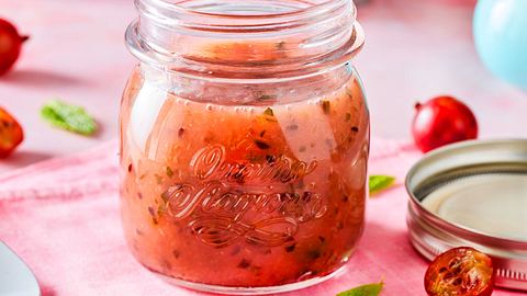 Ein offenes Glas mit Stachelbeermarmelade steht auf einem rosa Tuch, umgeben von halbierten Stachelbeeren und Minzblättern. - Foto: House of Food / Bauer Food Experts KG