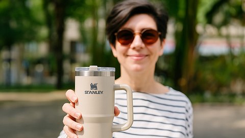 Lächelnde Frau mit Sonnenbrille hält beigen Stanley Cup in die Kamera. - Foto: iStock / DALL-E 3