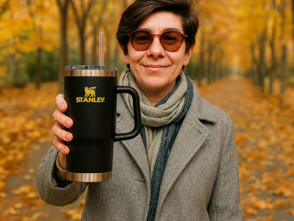 Eine Person mit Sonnenbrille und Schal hält einen Stanley Cup Becher in einer herbstlichen Allee in die Kamera.