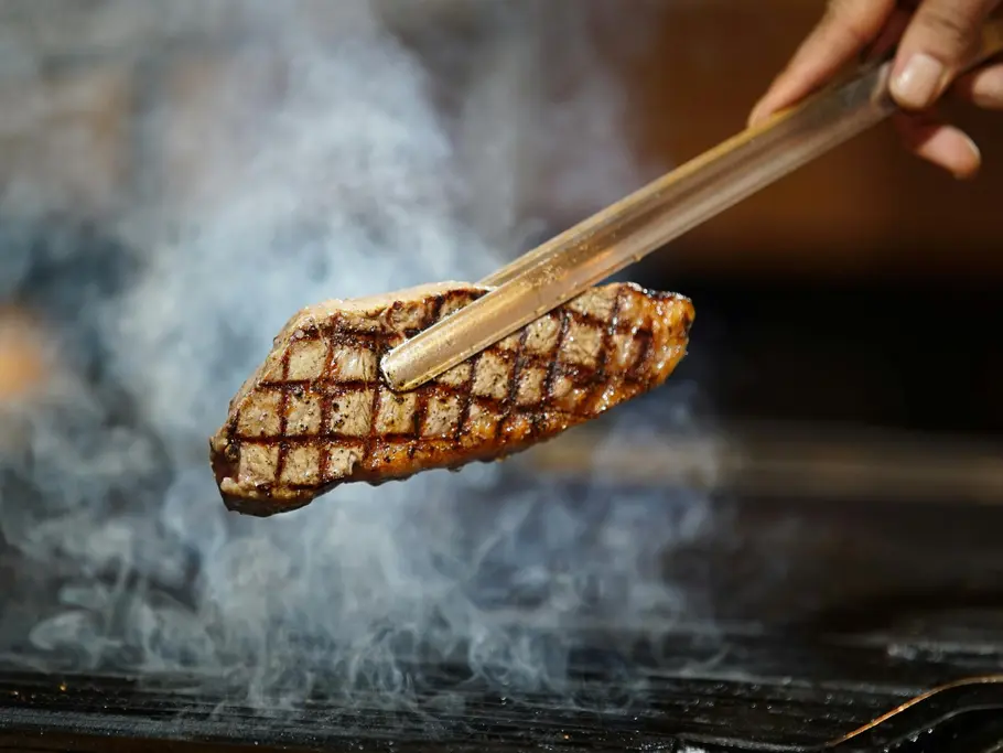 Ein gegrilltes Steak mit Rautenmuster wird von einer feinen Grillzange vom rauchenden Grillrost gehoben