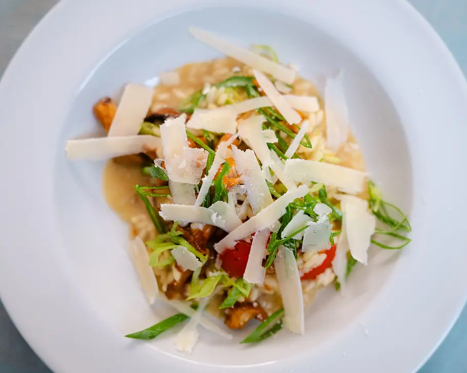 Ein Risotto mit Pfifferlingen, Kirschtomaten, Frühlingszwiebeln und Parmesanhobeln serviert in einem tiefen weißen Teller.