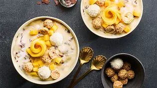 Zwei Schalen mit cremiger Stracciatella-Bowl, garniert mit Mangowürfeln, Mangoröschen, Kokoskugeln, kleinen Mandelgebäcken und Schokostückchen, serviert auf einem dunklen Untergrund. Neben den Schalen liegen goldene Löffel und eine kleine Schale mit Gebäck. - Foto: LECKER @ Bauer Media Group