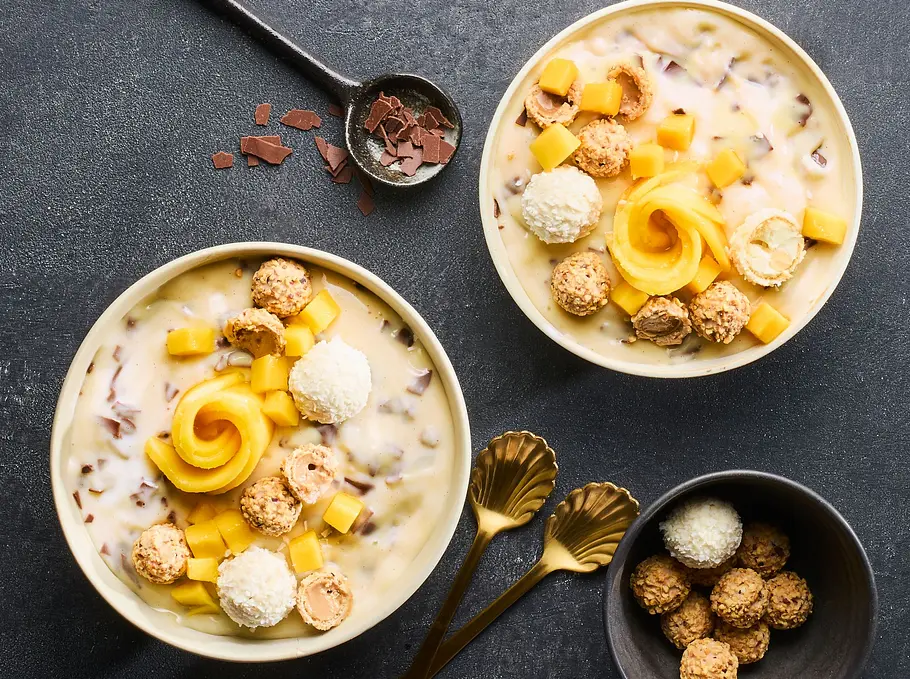 Zwei Schalen mit cremiger Stracciatella-Bowl, garniert mit Mangowürfeln, Mangoröschen, Kokoskugeln, kleinen Mandelgebäcken und Schokostückchen, serviert auf einem dunklen Untergrund. Neben den Schalen liegen goldene Löffel und eine kleine Schale mit Gebäck.