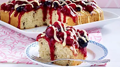 Stracciatella-Rührkuchen mit roter Grütze und Vanillesoße. Ein Stück befindet sich auf einem Teller, der ganze Kuchen im Hintergrund. - Foto: LECKER @ Bauer Media Group