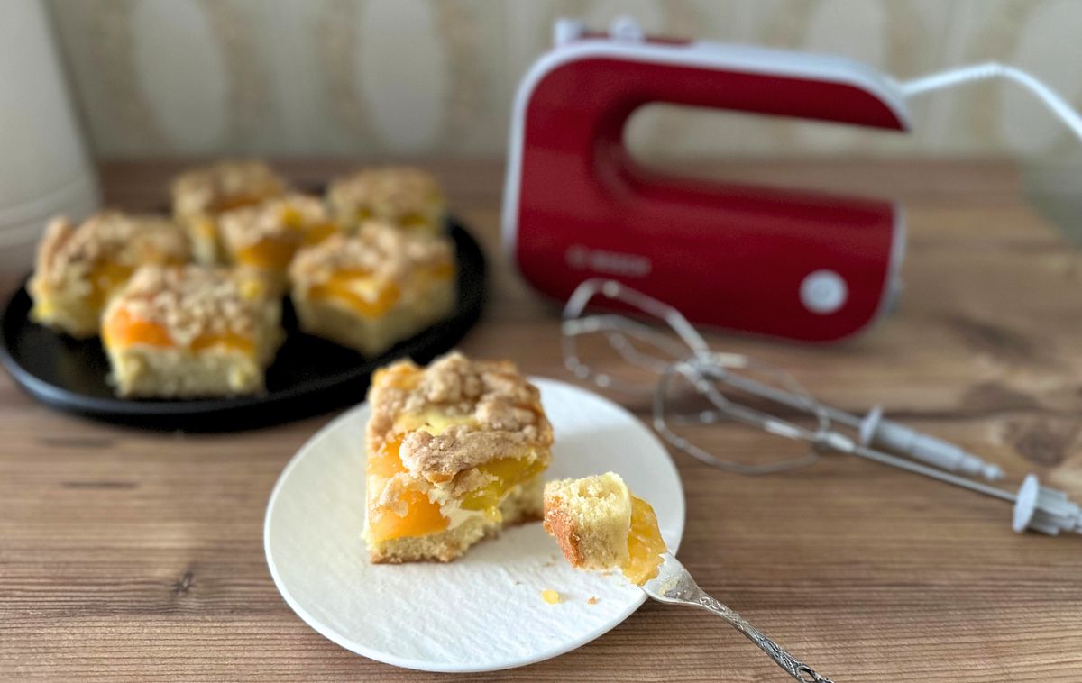 Ein angegabeltes Stück Aprikosen-Schmandkuchen mit Streuseln steht auf einer hölzernen Arbeitsfläche vor dem Bosch Styline Handrührgerät mit danebenliegenden Quirlen