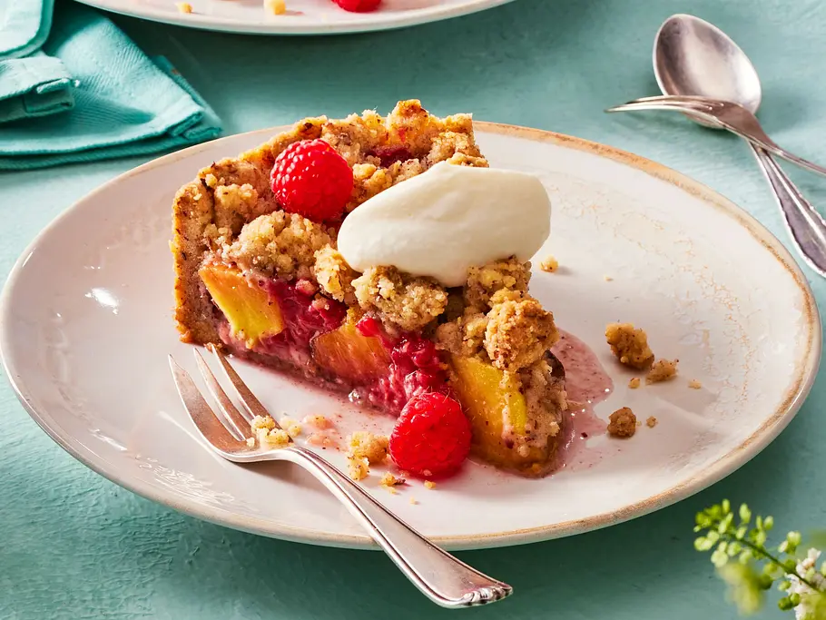 Zwei Stücke Streuselkuchen Pfirsich-Melba auf hellen Tellern mit frischen Pfirsichen, Himbeeren und Streuseln, jeweils mit einem Klecks Sahne, dazu Gabeln und ein Glas Kaffee auf mintfarbener Unterlage.