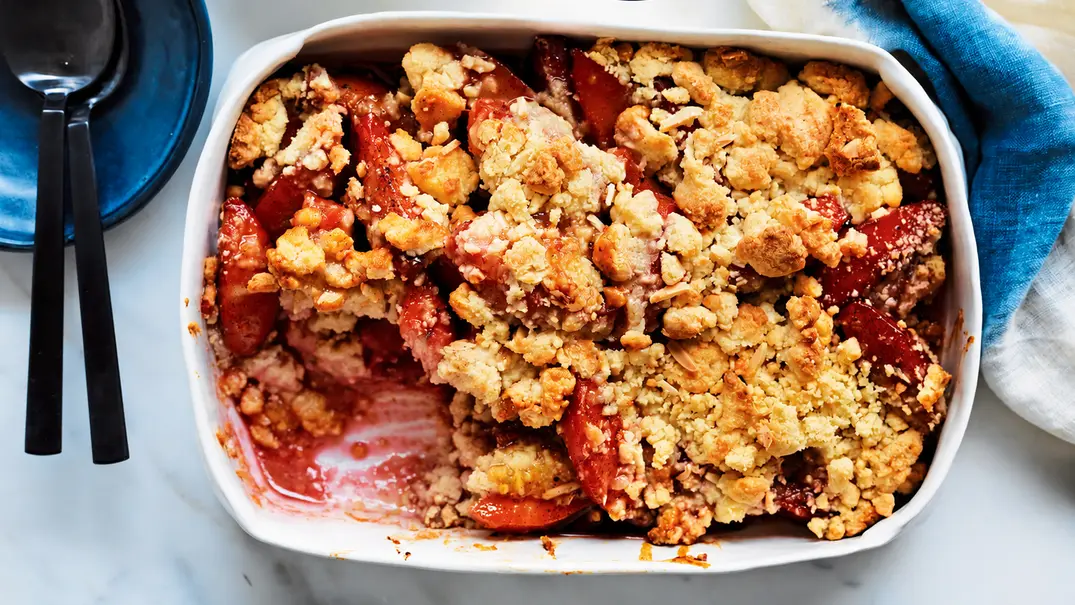 Ein angelöffelter Pflaumen-Crumble in einer weißen Auflaufform auf dem gedeckten Tisch. Links oben sind blaue Kuchenteller mit schwarzen Löffeln. - Foto: LECKER @ Bauer Media Group