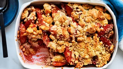 Ein angelöffelter Pflaumen-Crumble in einer weißen Auflaufform auf dem gedeckten Tisch. Links oben sind blaue Kuchenteller mit schwarzen Löffeln. - Foto: LECKER @ Bauer Media Group