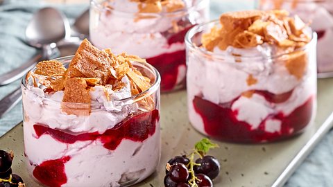 Vier Dessertgläser mit einer Schicht aus schwarzem Johannisbeerpüree und Sahne-Mascarpone-Creme, getoppt mit knusprigen Keksstücken, dekoriert mit frischen schwarzen Johannisbeeren auf einem Tablett. - Foto: House of Food / Bauer Food Experts