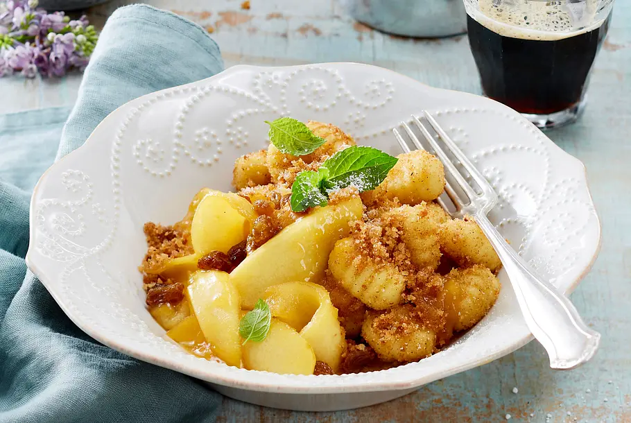 Weiße Schüssel mit süßen Gnocchi, Apfelspalten, Zimtbröseln und Minze garniert.