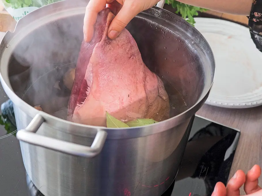 Ein Stück roher Tafelspitz wird in einen großen Topf mit heißer Brühe und Gewürzen gelegt, während Dampf aufsteigt.