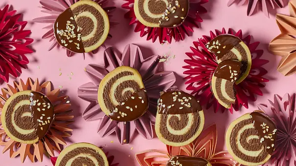Runde Tahin-Swirl-Taler mit dekorativem Wirbelmuster, zur Hälfte in Schokolade getaucht und mit Sesam bestreut, angerichtet auf bunten Stern-Unterlagen auf rosa Oberfläche. - Foto: LECKER @ Bauer Media Group