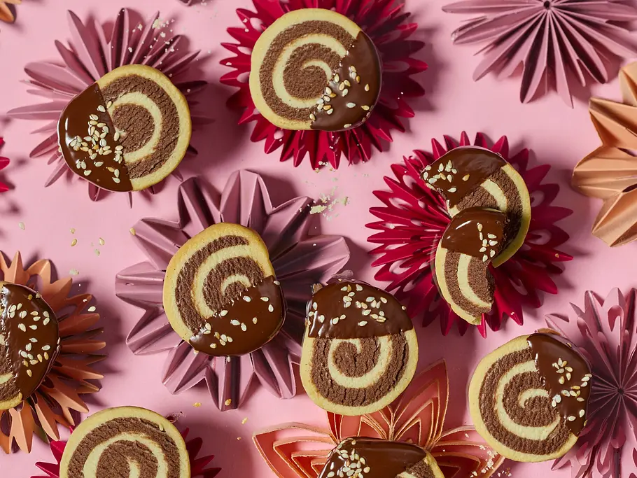 Runde Tahin-Swirl-Taler mit dekorativem Wirbelmuster, zur Hälfte in Schokolade getaucht und mit Sesam bestreut, angerichtet auf bunten Stern-Unterlagen auf rosa Oberfläche.