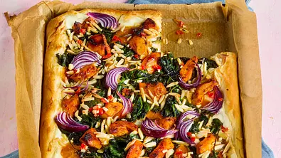 Rechteckige Pizza auf Backpapier mit Tandoori-Hähnchenstücken, roter Zwiebel, Spinat, Mandelsplittern und roten Chilistücken, serviert auf einem Backblech. Im Hintergrund Teller mit einem Pizzastück und ein Glas auf einem rosanen Untergrund. - Foto: StockFood Studios/Meike Bergmann Rechteckige Pizza auf Backpapier mit Tandoori-Hähnchenstücken, roter Zwiebel, Spinat, Mandelsplittern und roten Chilistücken, serviert auf einem Backblech. Im Hintergrund Teller mit einem Pizzastück und ein Glas auf einem rosanen Untergrund. - Foto: StockFood Studios/Meike Bergmann