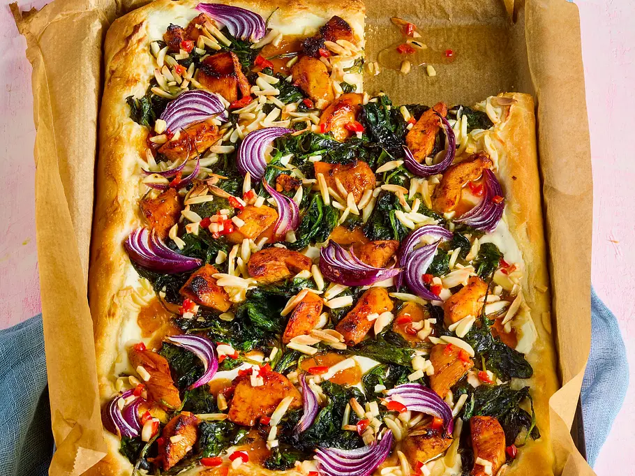 Tandoori-Chicken-Pizza Rechteckige Pizza auf Backpapier mit Tandoori-Hähnchenstücken, roter Zwiebel, Spinat, Mandelsplittern und roten Chilistücken, serviert auf einem Backblech. Im Hintergrund Teller mit einem Pizzastück und ein Glas auf einem rosanen Untergrund.
